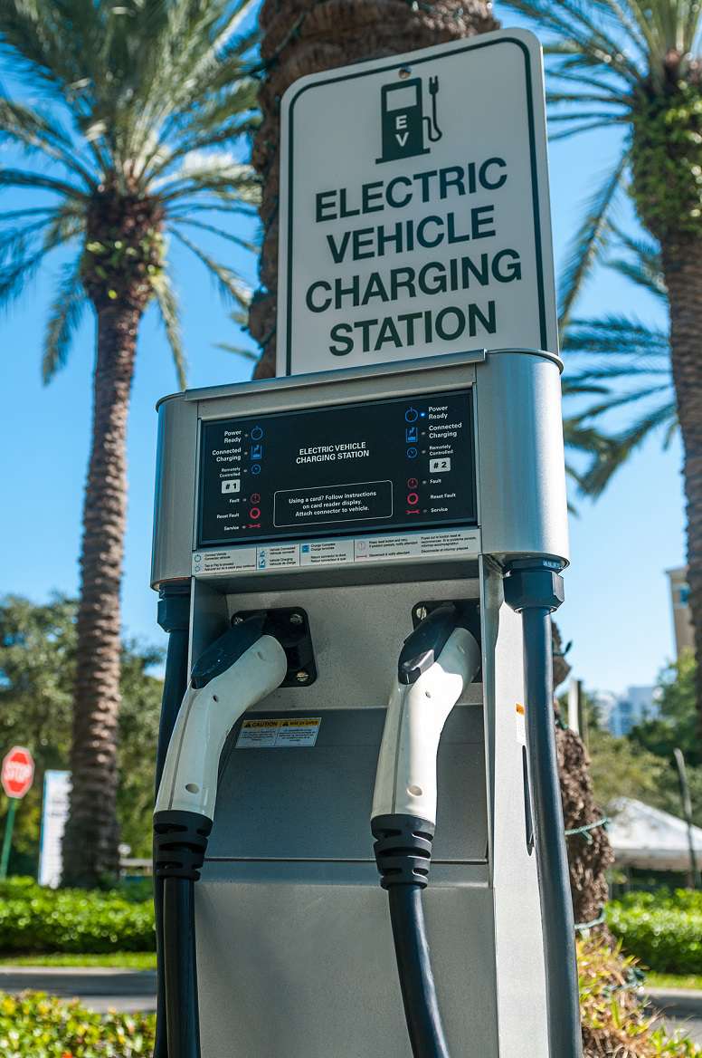 Electric vehicle charging station sign on top of a public charging station with palm trees in background.